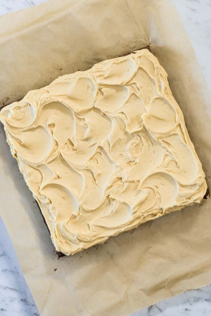 An overhead shot of a square chocolate brownie slab topped with a thick, decorative layer of tan peanut butter frosting, resting on a sheet of parchment paper.