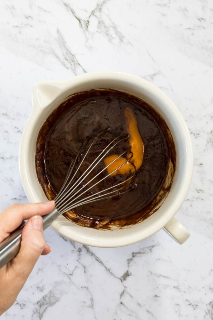 Metal whisk mixes a whole raw egg into thick, dark chocolate batter inside a white ceramic bowl for chocolate and peanut butter brownies.