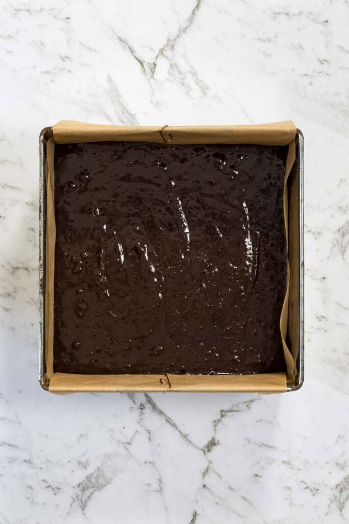 Glossy dark chocolate batter is smoothed out into an even layer inside a parchment-lined square baking pan for chocolate and peanut butter brownies.