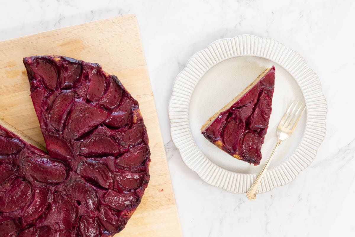 Slice of golden-brown upside down cake topped with deep purple cooked plums is served on a white ridged plate with a silver fork.