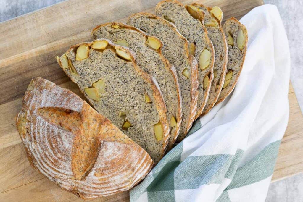 A whole, baked, and dusted loaf of potato and poppy seed bread and five thick slices resting on a wooden cutting board, with the slices tucked into a white and green striped tea towel.