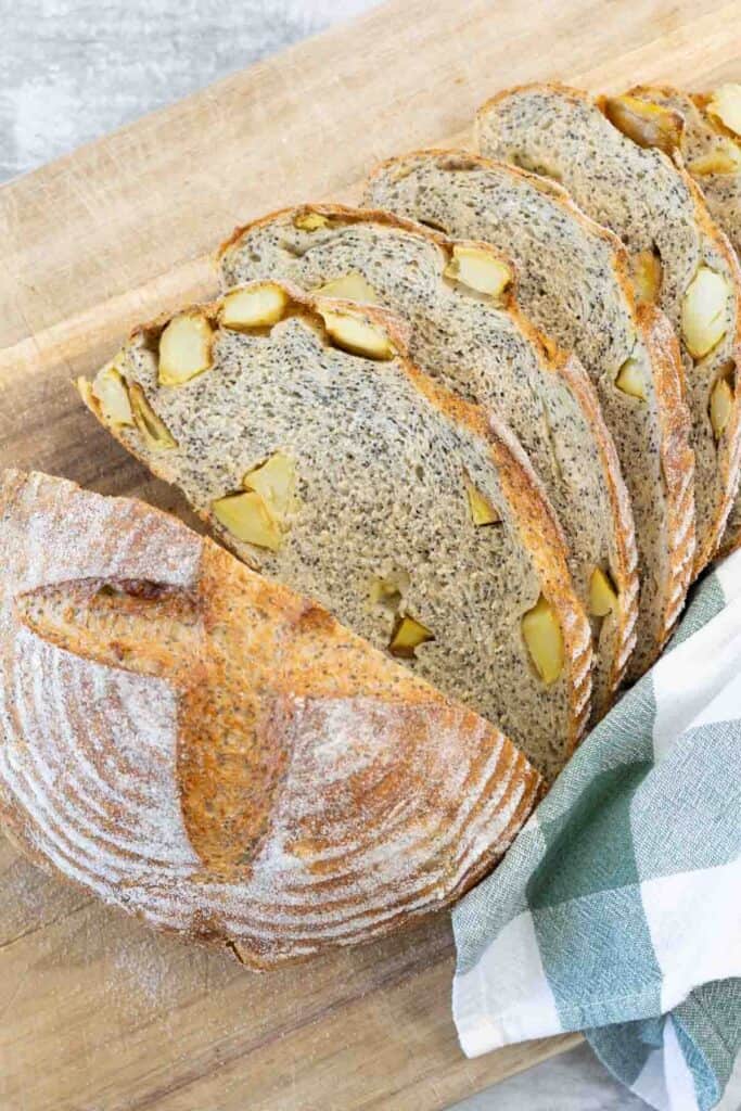 A fully baked and sliced loaf of crusty sourdough-style potato and poppy seed bread resting on a wooden board, revealing an interior cross-section dotted with black poppy seeds and yellow chunks of roasted potato.
