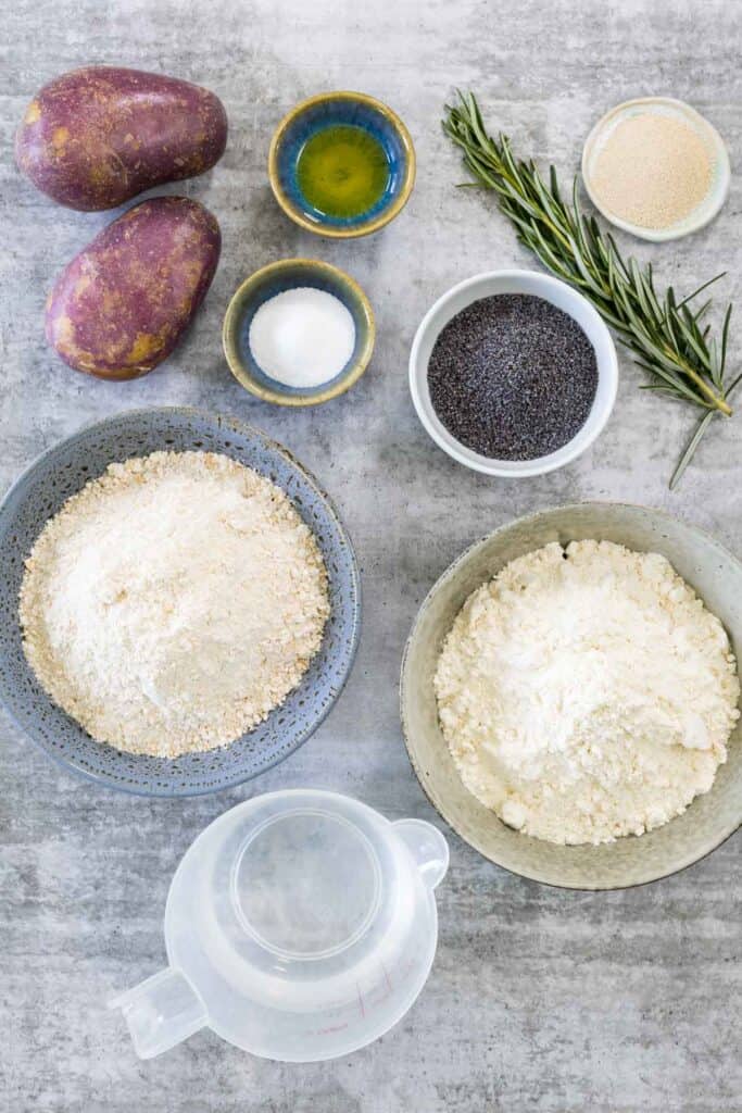 Ingredients for potato and poppy seed bread arranged on a gray surface, including potatoes, flour, poppy seeds, rosemary, yeast, olive oil, salt, and water.