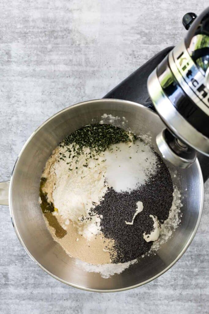 Flour, yeast, poppy seeds, salt, and chopped rosemary in a stand mixer bowl before kneading dough for potato and poppy seed bread.