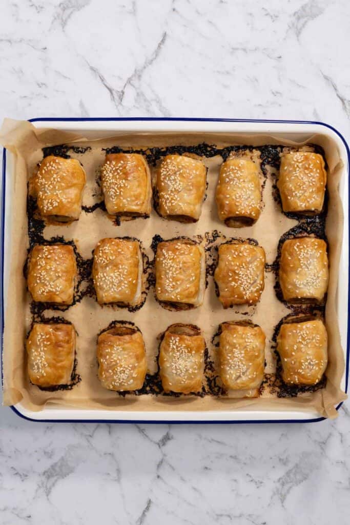 An overhead view of fifteen freshly baked pork and apple sausage rolls on parchment paper inside a blue-rimmed baking pan, featuring puffed, golden-brown flaky pastry topped with toasted white sesame seeds for sausage rolls.