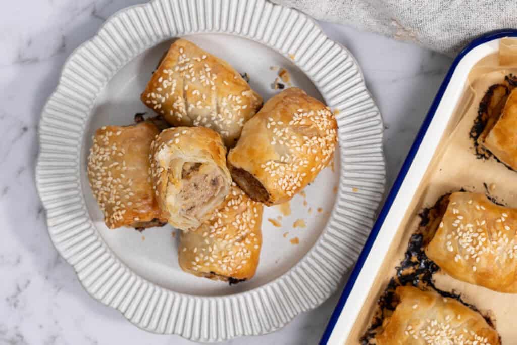 A close-up, overhead shot of five golden-brown pork and apple pastry rolls piled onto a ribbed white ceramic plate next to a blue-rimmed baking pan, with one roll cut in half to show the cooked meat and apple filling inside for sausage rolls.