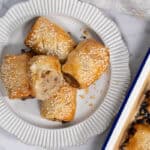 A close-up, overhead shot of five golden-brown pork and apple pastry rolls piled onto a ribbed white ceramic plate next to a blue-rimmed baking pan, with one roll cut in half to show the cooked meat and apple filling inside for sausage rolls.