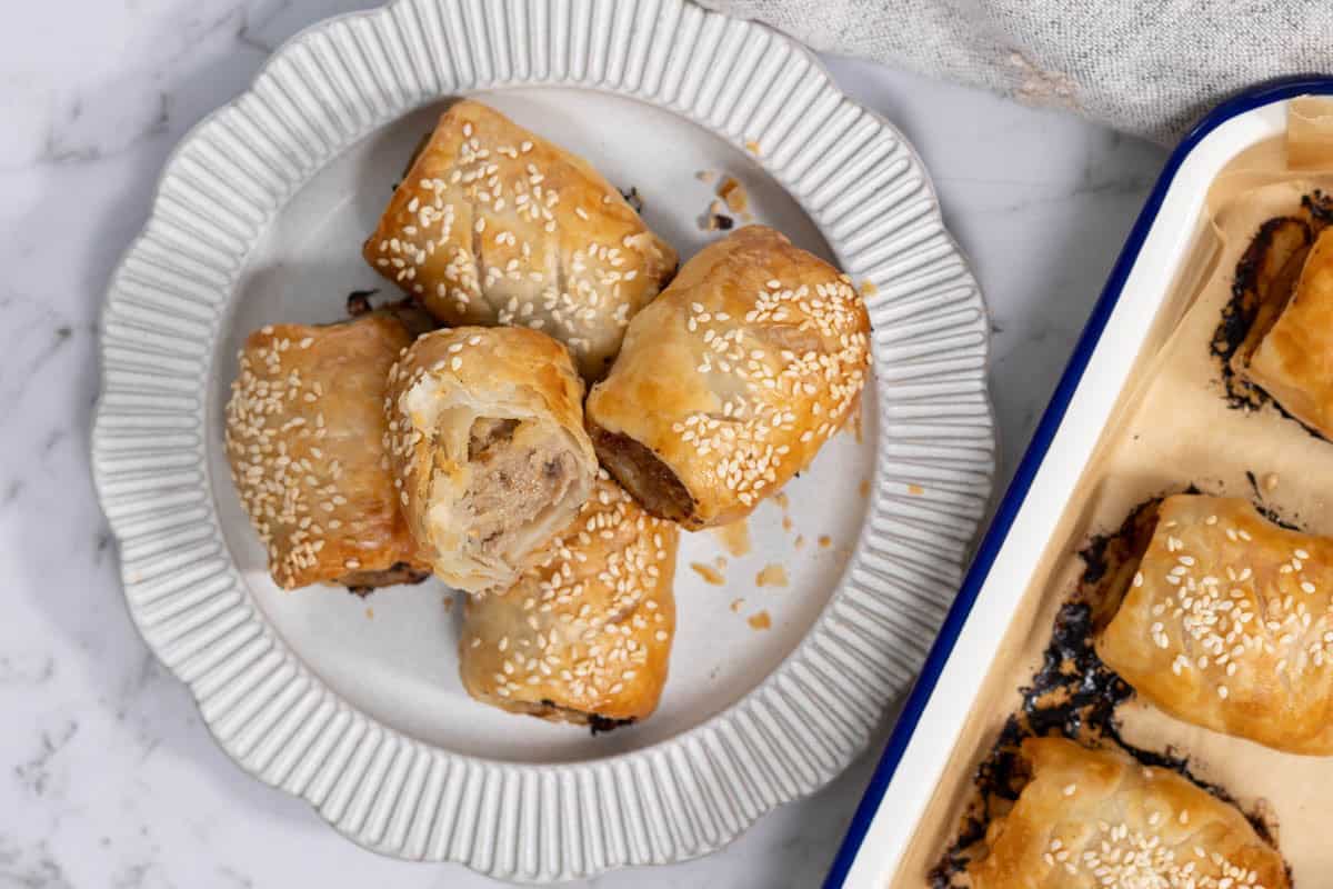 A close-up, overhead shot of five golden-brown pork and apple pastry rolls piled onto a ribbed white ceramic plate next to a blue-rimmed baking pan, with one roll cut in half to show the cooked meat and apple filling inside for sausage rolls.