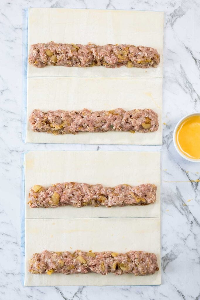 Four long logs of ground pork and apple filling are placed down the center of cut puff pastry sheets on a marble surface next to a small bowl of egg wash for sausage rolls.