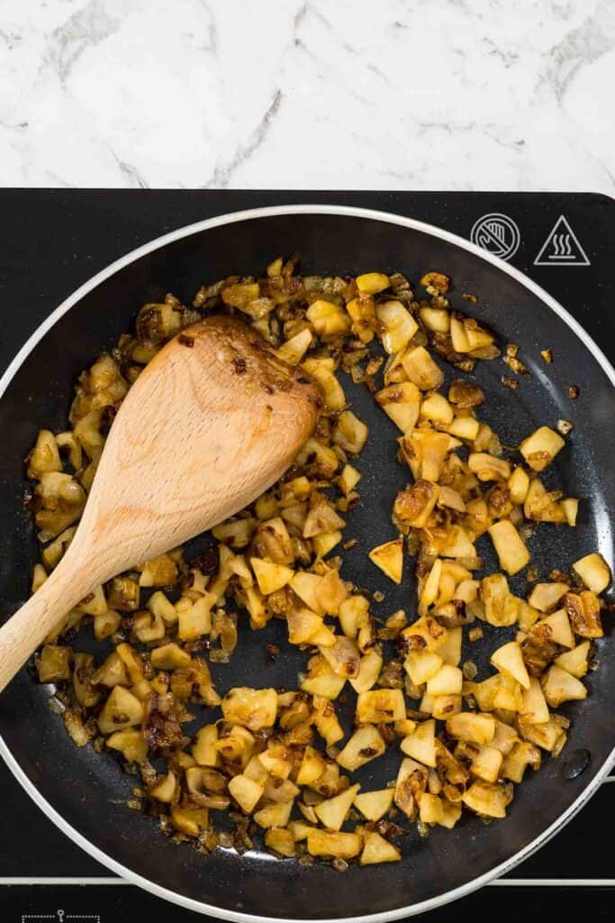 Diced green apples and onions are deeply caramelized and tender in a black skillet, being stirred with a wooden spatula for sausage rolls.