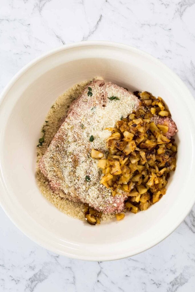 Raw ground pork, panko breadcrumbs, seasonings, fresh herbs, and the cooked caramelized apple and onion mixture are combined together inside a large white mixing bowl for sausage rolls.
