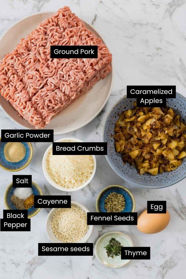 An overhead labeled view of filling ingredients on a marble surface, featuring Ground Pork, Caramelized Apples, Bread Crumbs, Garlic Powder, Salt, Black Pepper, Cayenne, Fennel Seeds, Sesame seeds, Thyme, and an Egg for sausage rolls.