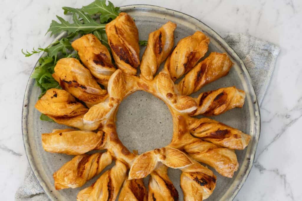 A red pesto and puff pastry wreath, made by twisting strips of pastry attached to a central ring, then baking. The wreath is served on a gray ceramic plate with salad leaves for garnish.