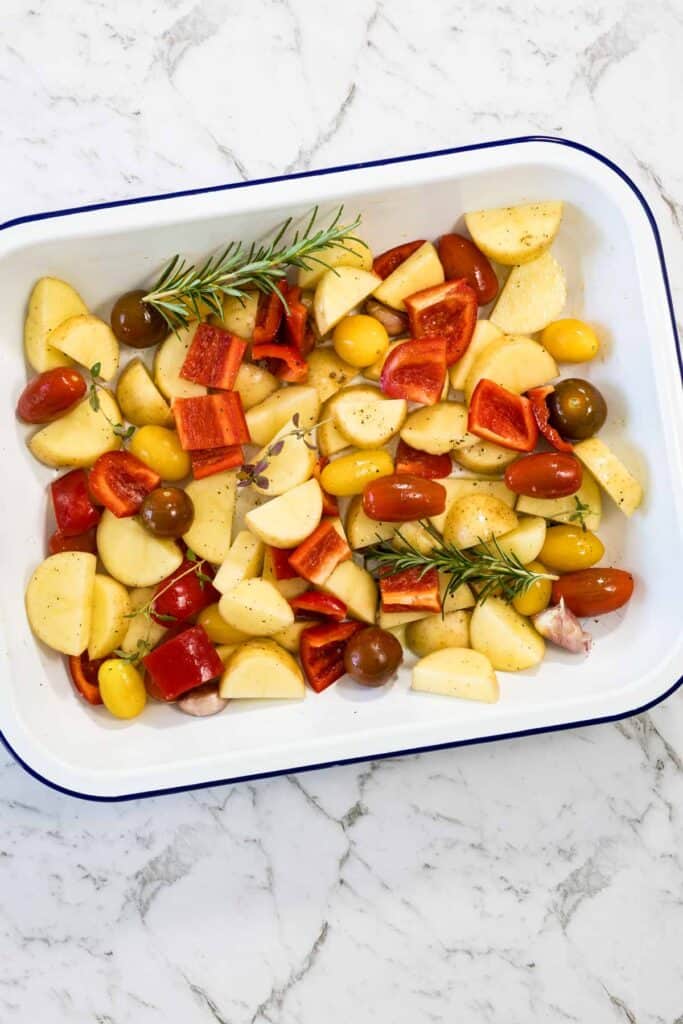 Diced potatoes, chopped red bell pepper, colorful cherry tomatoes, garlic cloves, and fresh rosemary sprigs are seasoned and arranged in a white baking dish with a blue rim for a sausage bake.
