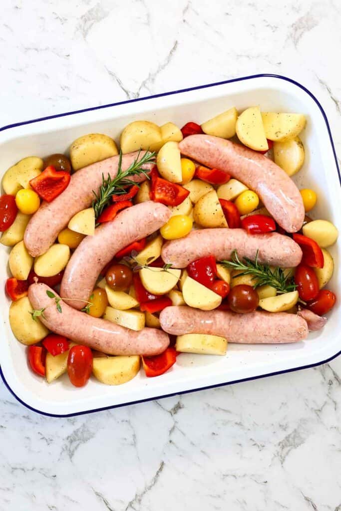 Raw pork sausages are nestled into the bed of seasoned chopped potatoes, red bell peppers, cherry tomatoes, and rosemary herbs in a white baking pan for a sausage bake.