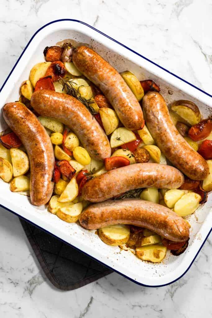 Cooked, browned pork sausages lie across roasted, golden-edged potatoes, softened red bell peppers, and blistered tomatoes in a white enamel baking dish for a sausage bake.