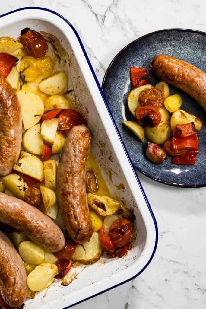 A portion of juicy roasted sausage, golden potatoes, blistered cherry tomatoes, and red peppers is served on a dark blue ceramic plate next to the main enamel baking dish for a sausage bake.