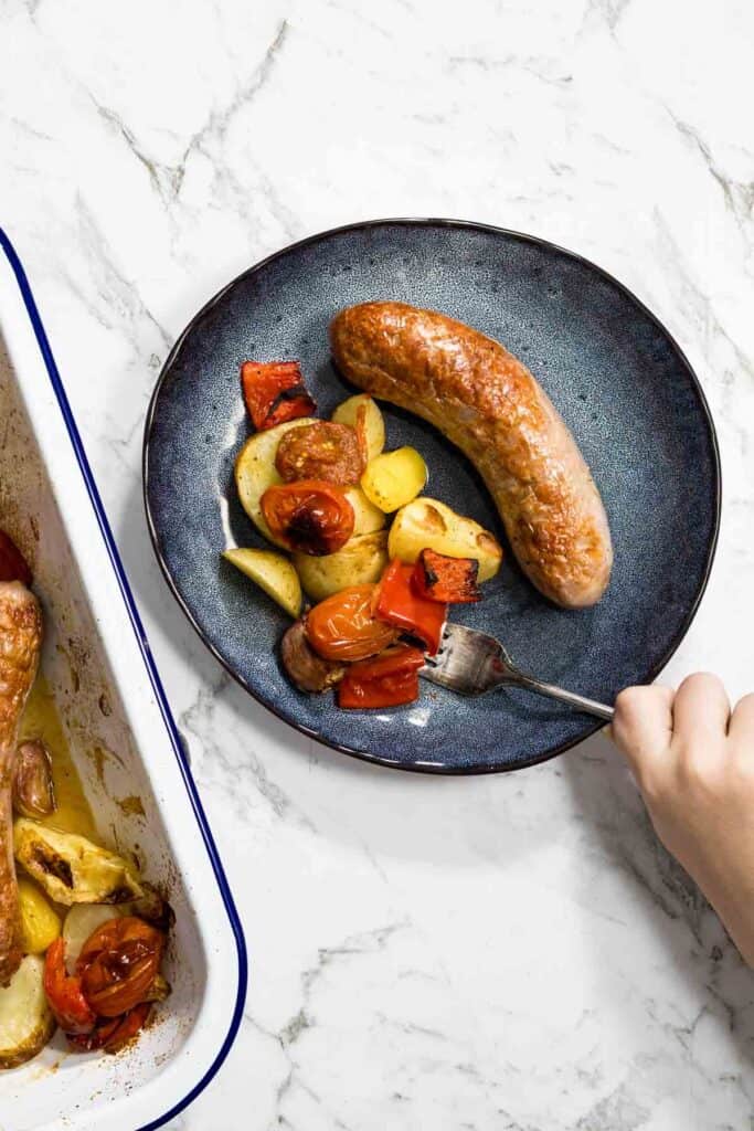 A hand holds a silver fork, picking up a piece of roasted red pepper from a dark blue plate containing a golden-brown sausage, roasted potatoes, and blistered tomatoes for a sausage bake.