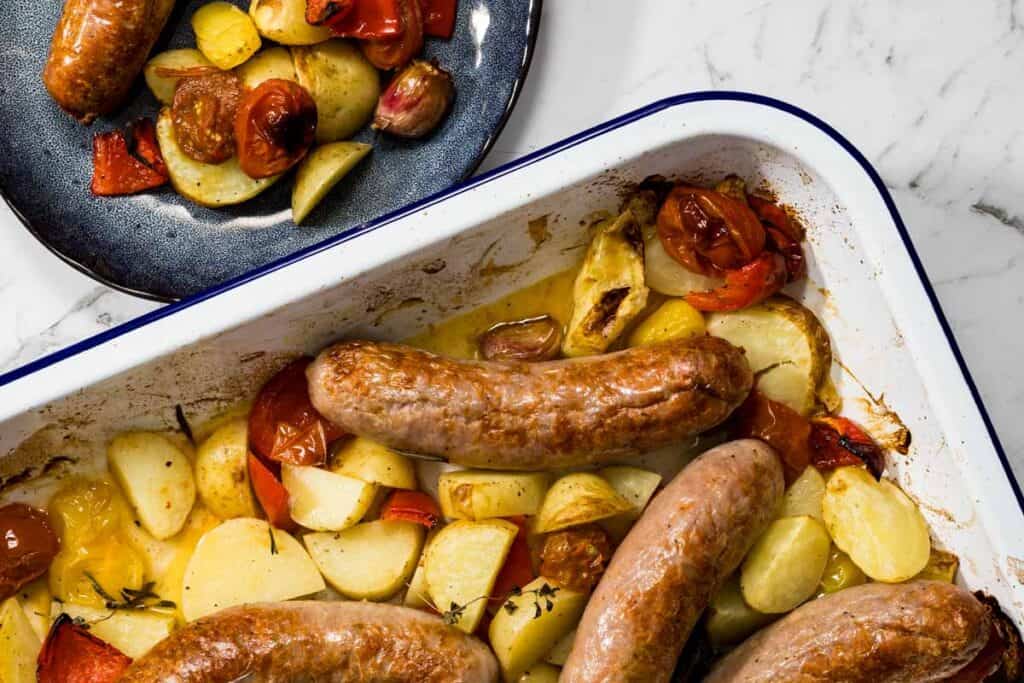 Overhead landscape view of a white baking dish containing roasted sausages, golden potatoes, and blistered tomatoes next to a dark blue plate with a served portion for a sausage bake.