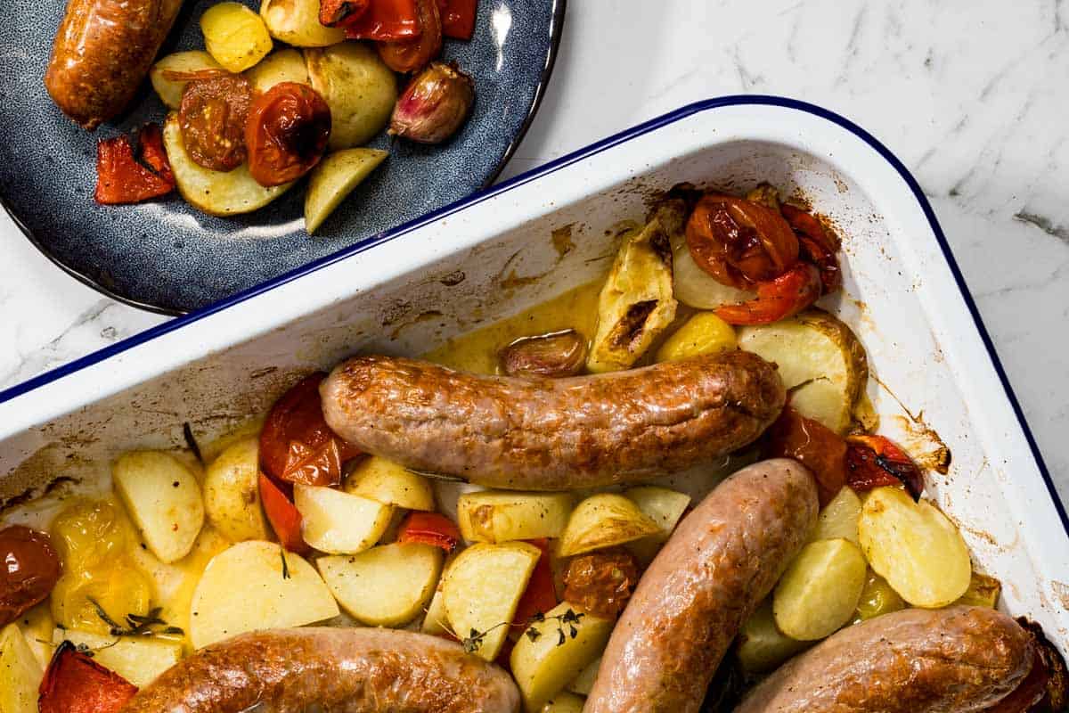 Overhead landscape view of a white baking dish containing roasted sausages, golden potatoes, and blistered tomatoes next to a dark blue plate with a served portion for a sausage bake.