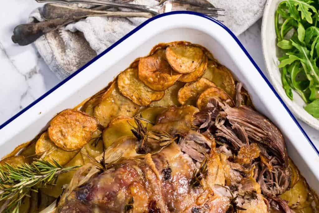A close-up, angled view of roasted lamb leg with tender meat falling away from the bone, surrounded by golden-brown crispy potato slices in a white enamel pan.