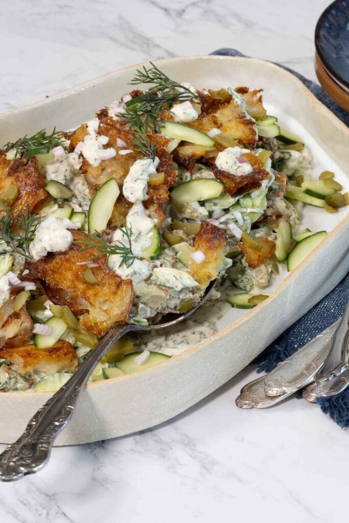 An organic-shaped white platter with smashed potato salad on it. In the salad you can see crispy potatoes, a creamy dressing, cucumbers, pickles and herbs. 