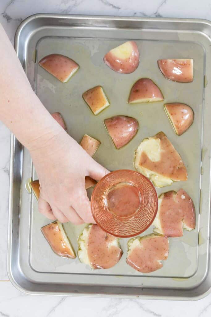 A hand holds a glass, using the base of the glass to press down on steamed potatoes to squash them, ready for roasting.