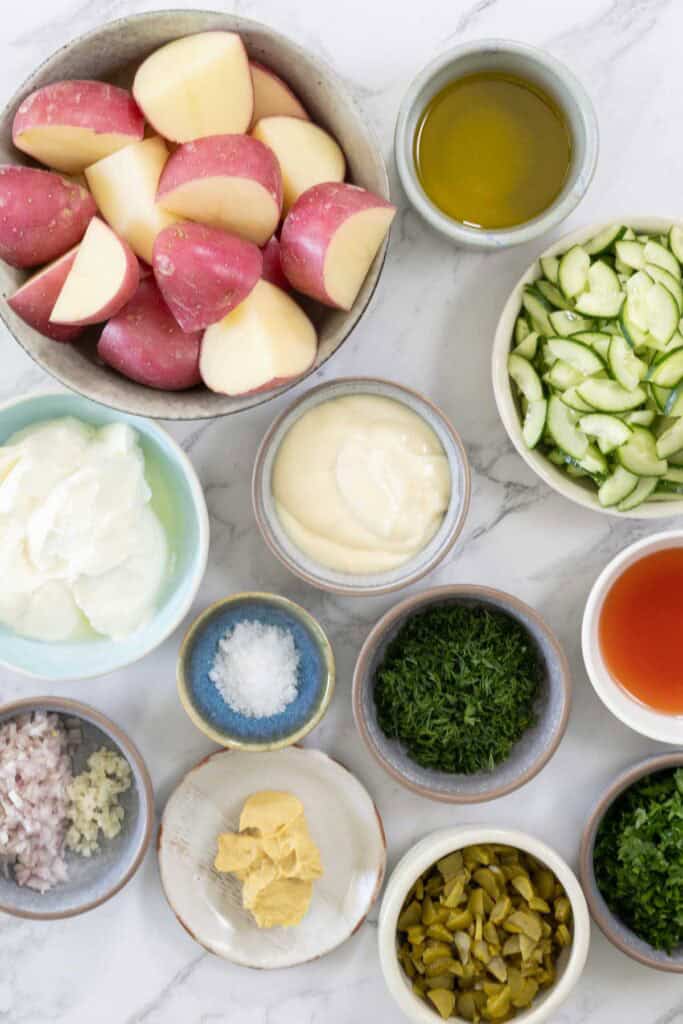 A top-down view of the ingredients for smashed potato salad. Potatoes, olive oil, cucumber, vinegar, herbs, mayonnaise, yoghurt, shallots, mustard, pickles and salt. 