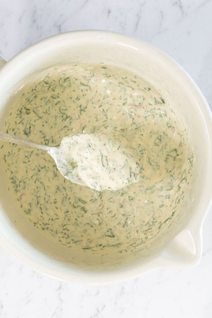 A large ceramic bowl, viewed from the top down. In the bowl is freshly mixed dressing for smashed potato salad. It looks creamy and thick, and is flecked with herbs. 