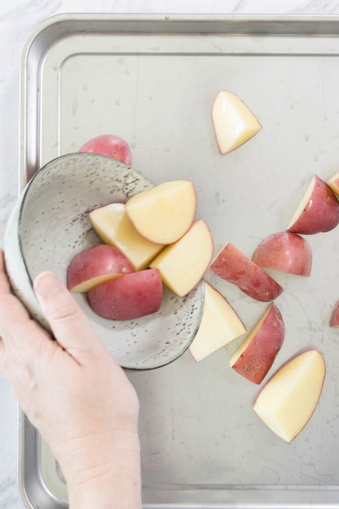 A hand tips a bowl full of diced red-skinned potatoes onto a metal tray ready for steaming. 