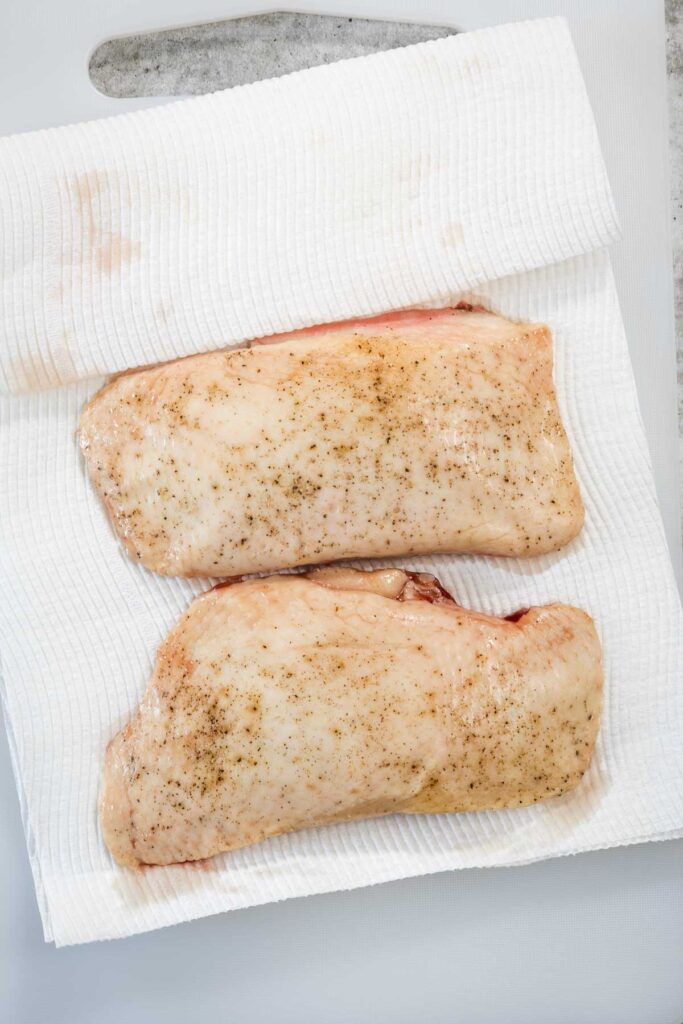 Two duck breasts on a white chopping board. The breasts are being dried with paper towels after being cooked sous vide.