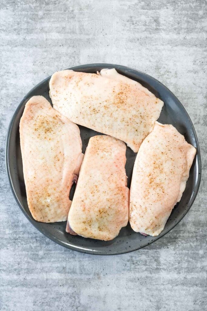A dark gray plate contains four raw duck breasts. The skin is visible on the breasts, and has been rubbed with salt and pepper for flavor.