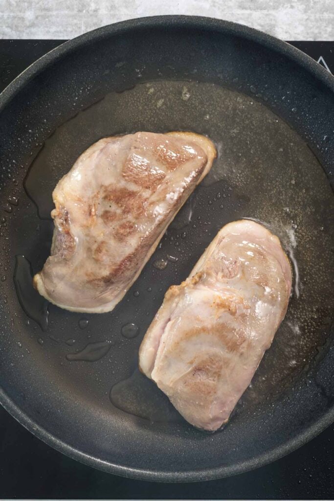 Two duck breasts, skin side down, in a hot frying pan. Fat can be seen pooling around the duck breasts; they are being browned and crisped after sous vide cooking.