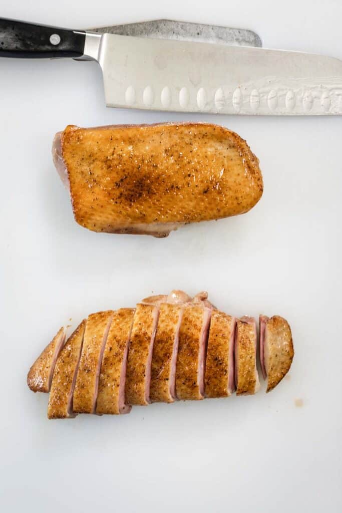 Two crispy skinned sous vide duck breasts on a white plastic chopping board. A large knife is visible at the top of frame, and one of the breasts has been sliced for serving.