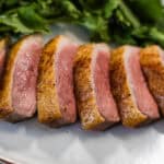 A close-up image of sliced crispy skinned sous vide duck breast, served on a white plate with cilantro leaves in the background.