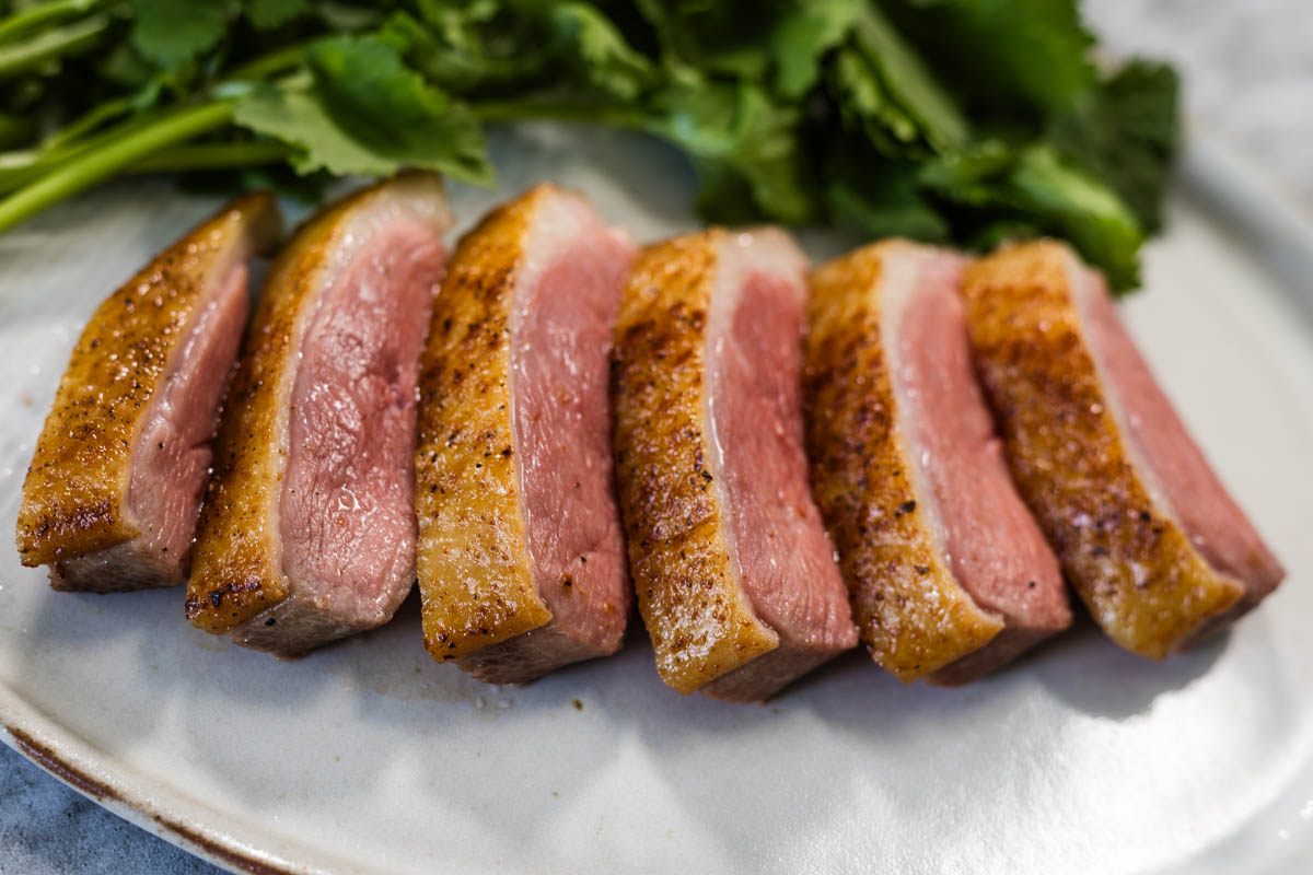 A close-up image of sliced crispy skinned sous vide duck breast, served on a white plate with cilantro leaves in the background.