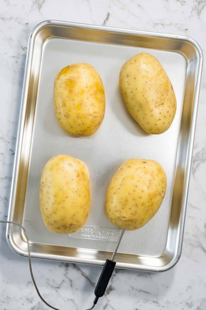 Four large yellow potatoes are arranged on a silver metal baking sheet, with a digital meat thermometer probe inserted into the bottom-right potato for steam baked potatoes.