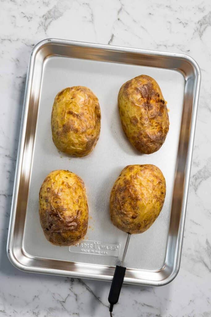 Four baked yellow potatoes with wrinkled, golden-brown crispy skins sit on a silver baking pan with a temperature probe still inserted into one potato for steam baked potatoes.