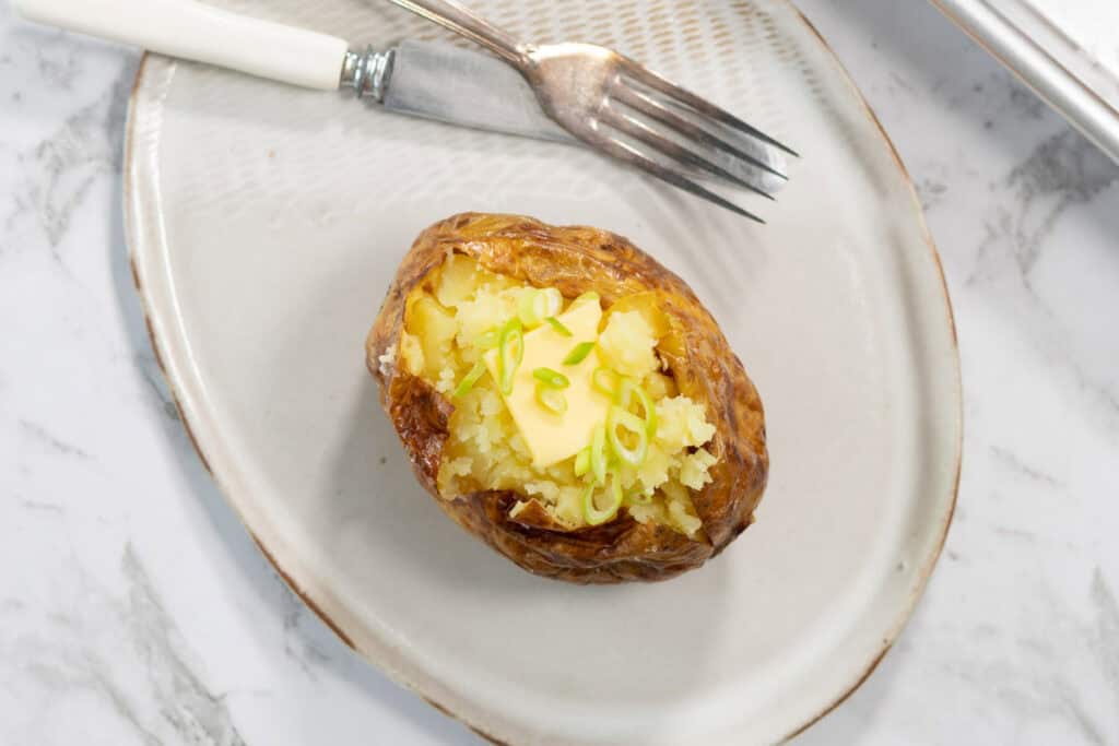 A single steam baked potato is split open on a light ceramic plate, revealing a fluffy yellow interior topped with a square melting pat of butter and sliced green onions next to a silver fork and knife.