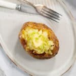 A single steam baked potato is split open on a light ceramic plate, revealing a fluffy yellow interior topped with a square melting pat of butter and sliced green onions next to a silver fork and knife.