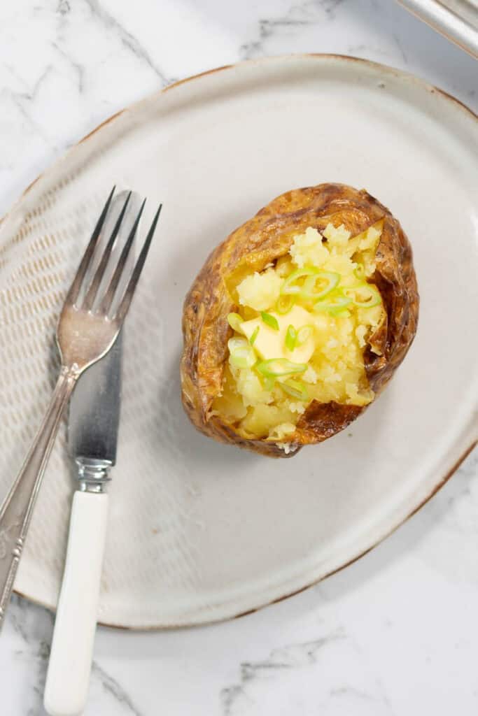 A close-up, top-down view of a split-open steam baked potato on an oval ceramic platter, showcasing the melting pat of butter and scattered sliced scallions alongside a vintage-style fork and knife.