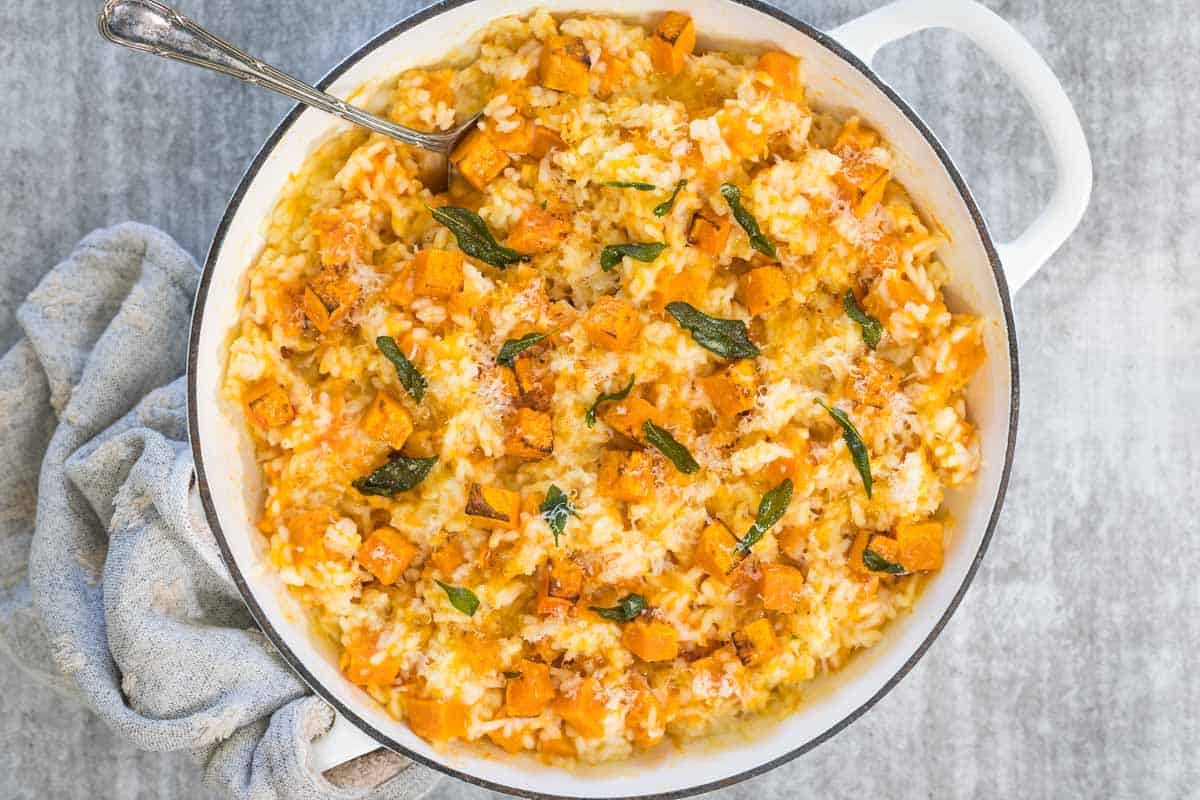 Pumpkin and crispy sage steamed risotto, served in a white pan and viewed from above. A gray cloth is wrapped around one handle of the pan and a silver spoon rests in the dish.