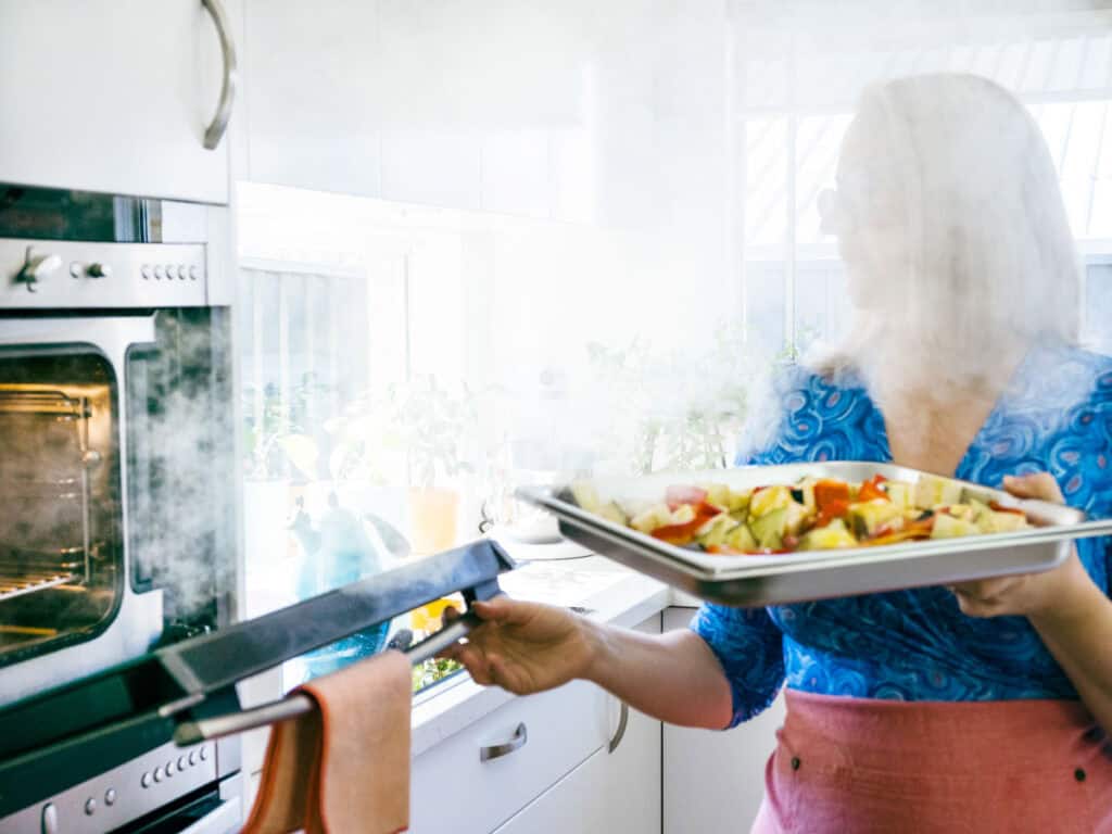 A woman holds a tray of vegetables. She is standing in front of an open steam oven, with steam billowing out from the inside of the oven. 