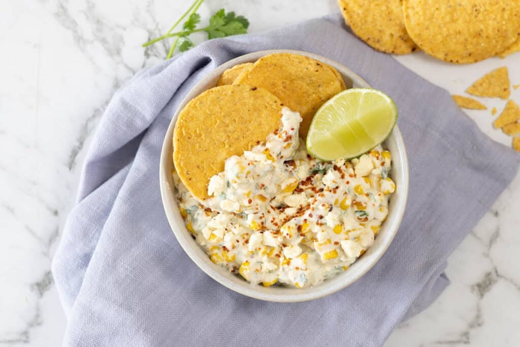 creamy corn appetizer topped with crumbled cheese, chili seasoning, and a fresh lime wedge, served with round tortilla chips in a ceramic bowl over a blue linen napkin