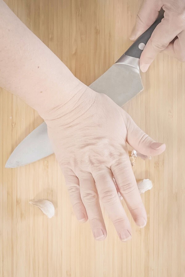 Crushing garlic cloves with the side of a knife on a wooden cutting board.