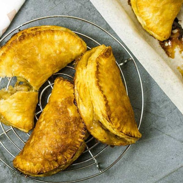 a gray benchtop with a round cake rack. On the rack are three puff pastry apple turnovers; one is broken open to show the apple filling inside.