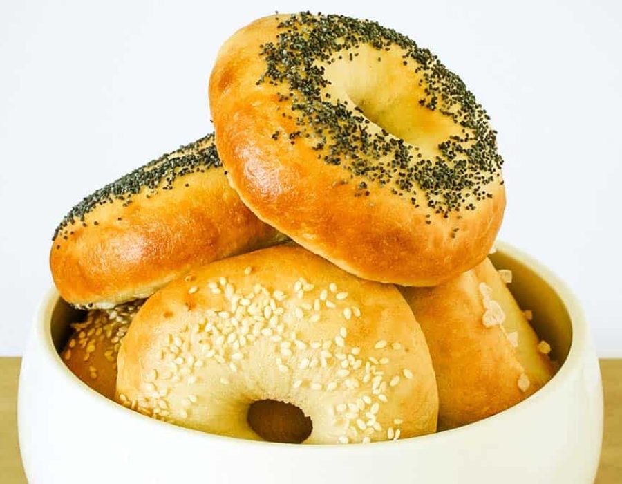 A white bowl with bagels, each topped with sesame seeds or poppy seeds or flaky salt, arranged in a pile
