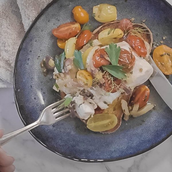 a blue plate with a piece of garlic toast, topped with flaky white fish, roasted cherry tomatoes, onions and fennel. A knife and fork are cutting the fish for eating.