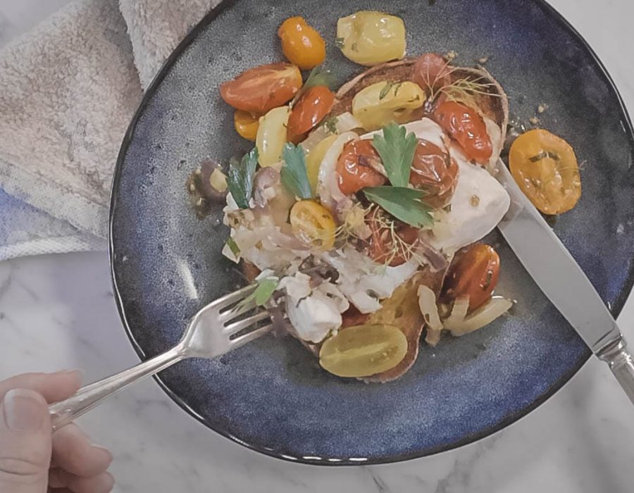 a blue plate with a piece of garlic toast, topped with flaky white fish, roasted cherry tomatoes, onions and fennel. A knife and fork are cutting the fish for eating.