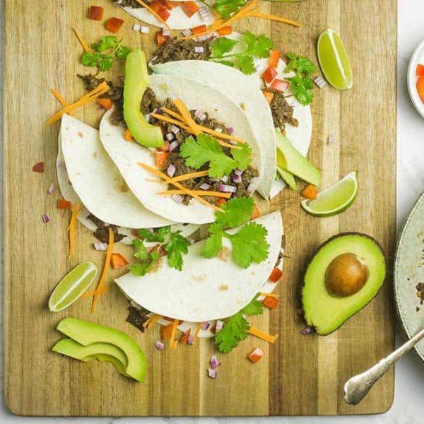 Close up view of soft tacos with shredded beef, julienned carrot, diced onion & tomato on a wooden board on a marble countertop.